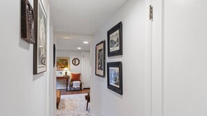 A hallway with framed pictures on the walls, a door at the end, and a small table with a lamp and a chair in the background.