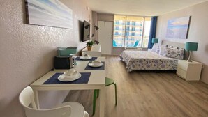 Habitación de hotel con cama, escritorio con silla, pequeña mesa con otra silla, televisor montado en la pared y ventana con vista a edificios.