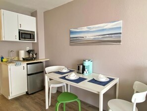 Una cocina compacta con armarios blancos, un microondas y una cafetera. Hay una mesa de comedor con cuatro sillas y un taburete verde. Un cuadro con una escena de playa adorna la pared.