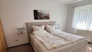 A bedroom with a bed, a bedside table, a nightstand, a wall-mounted picture, and a window with curtains.
