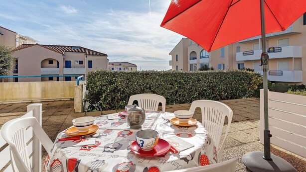 Een buitenterras met een tafel voor vier personen, omringd door witte stoelen, onder een rode parasol.