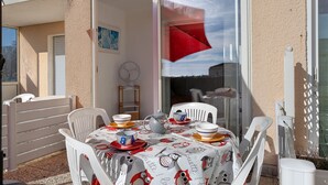 Een terras met een eettafel, buitenstoelen, een wit hekwerk en een raam met een rood luifeltje.