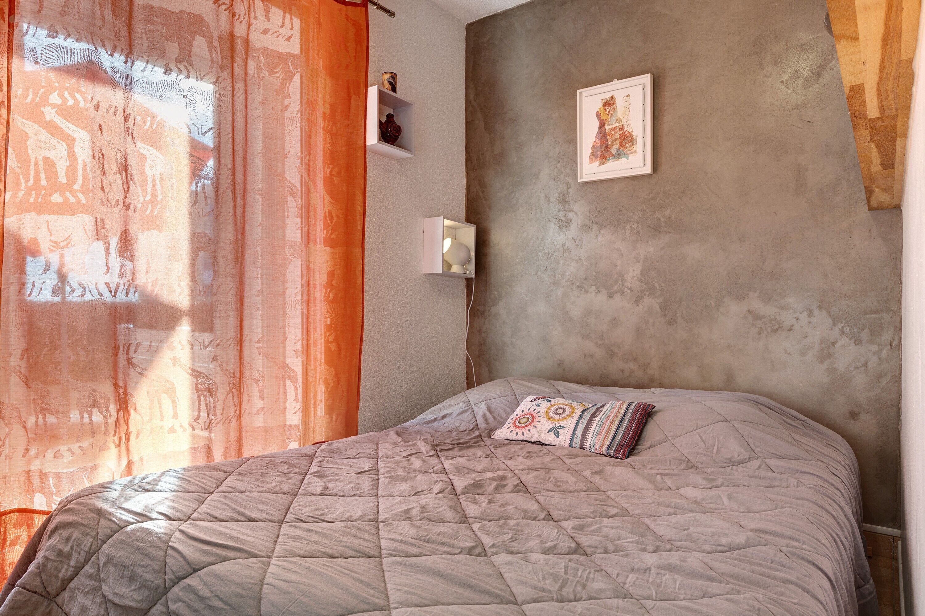Une chambre à coucher avec un lit, une étagère fixée au mur, un tableau encadré et une fenêtre avec des rideaux.