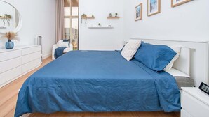 A bedroom with a large bed, a dresser, a chair, and a bedside table. There are wall-mounted shelves with plants and framed pictures.
