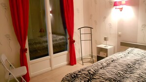 Un dormitori amb un llit, una cadira, una tauleta de nit, una làmpada, una finestra amb cortines i una làmpada penjada a la paret.