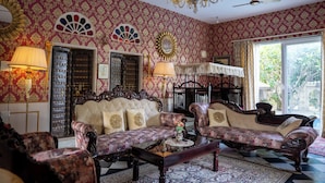 A room with patterned wallpaper, a chandelier, a ceiling fan, and a large window with a view. - Bhanpur Haveli | Hidden Luxury Haveli Amid Greenery & Peace (Jamwa Ramgarh)