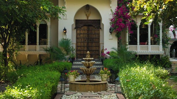 A courtyard with a fountain, a large wooden door, and a white balcony with intricate railings. - Bhanpur Haveli | Hidden Luxury Haveli Amid Greenery & Peace (Jamwa Ramgarh)