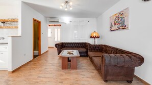 Ein Wohnzimmer mit braunem Ledersofa, einem Holztisch und einer Lampe. An der Wand hängt ein Gemälde und eine Tür führt in einen anderen Raum.