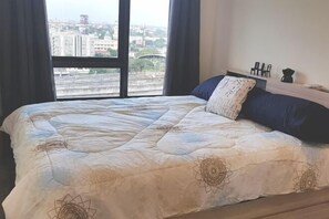 Un dormitorio con una cama grande, vistas de la ciudad a través de una ventana y una mesita de noche con lámpara.