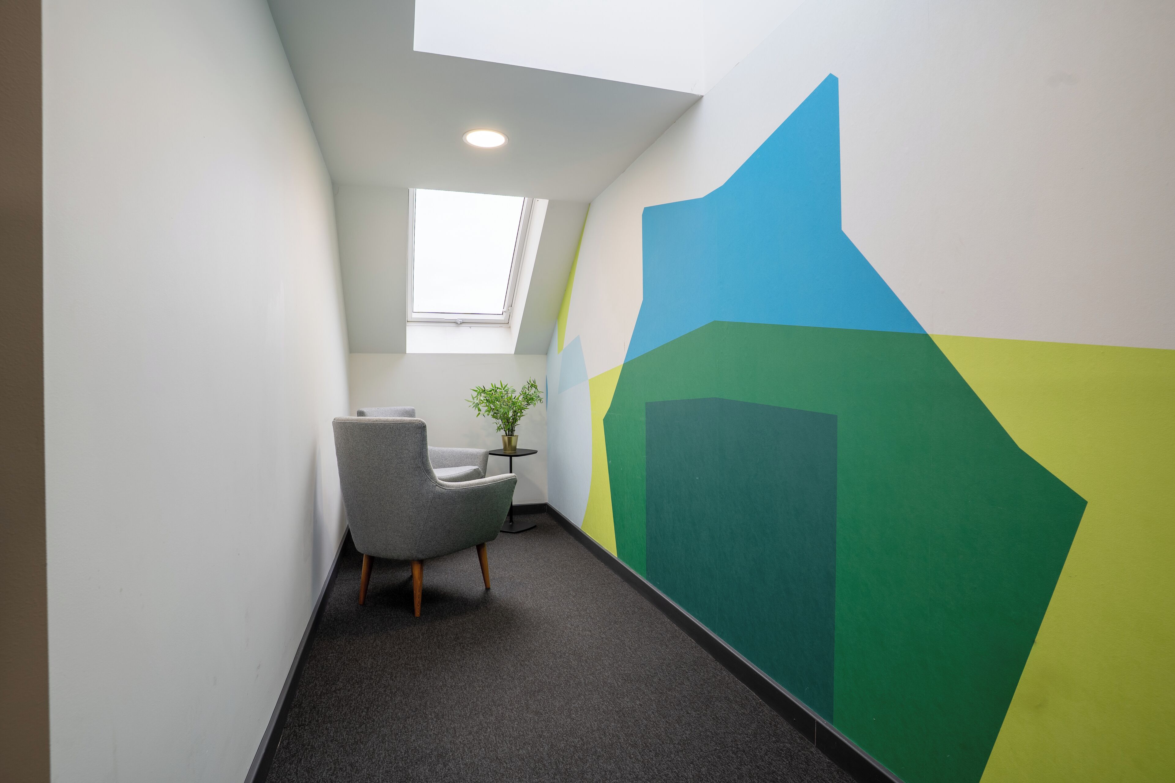 Un espace de bureau moderne avec une fresque murale aux motifs géométriques, un fauteuil de bureau gris, une petite table avec une plante et une lucarne.