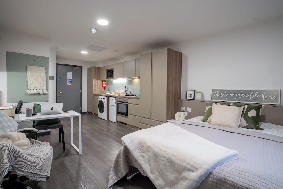 Une chambre d’hôtel moderne et compacte, dotée d’une kitchenette, d’un lit avec une grande fenêtre et d’un bureau avec un ordinateur portable.