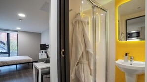 Um banheiro moderno com chuveiro de vidro, pia com parede de destaque amarela, e uma cama no quarto vizinho.