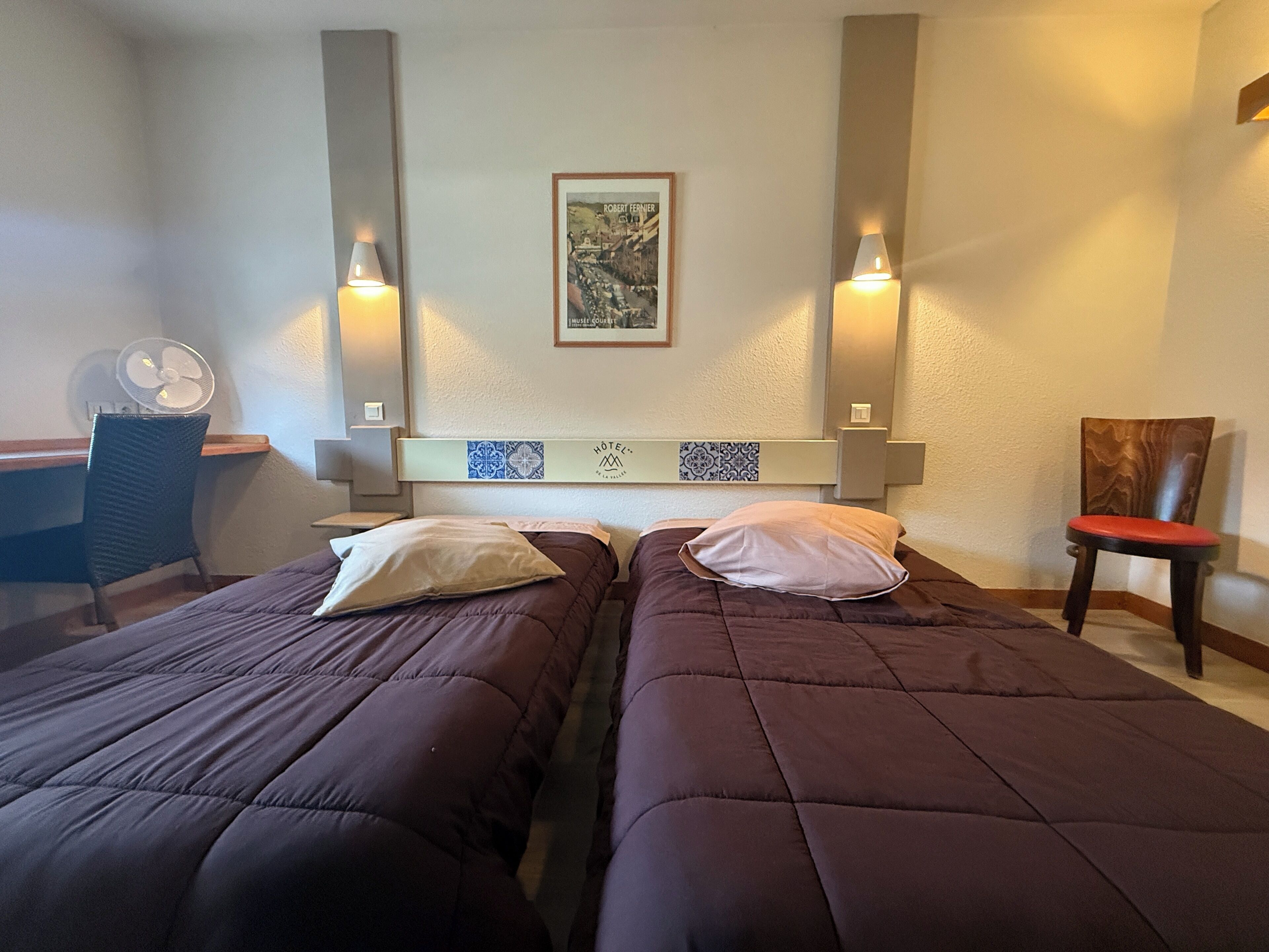 A hotel room with two beds, a desk, a chair, and a wall-mounted fan.