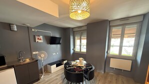 A modern kitchen with a dining area, grey walls, wooden flooring, a TV, and a window with blinds. - Hôtel de la Vallée (Ornans)
