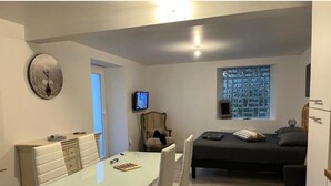 A compact living space with a dining area, a bed, a television, and a window with a view of a cityscape. - Hôtel de la Vallée (Ornans)