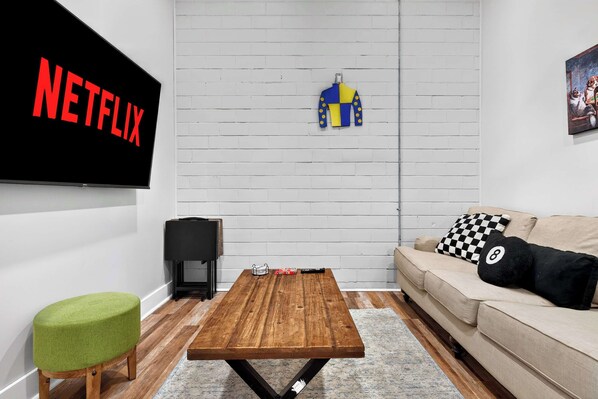 A living room with a TV displaying 'NETFLIX,' a wooden coffee table, a green ottoman, a beige sofa, and a checkered pillow. - The Billy Walters Suite | Modern Brick Retreat! (Louisville)