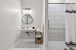 A modern bathroom with a white sink, round mirror, and a shower with a white curtain. - The Billy Walters Suite | Modern Brick Retreat! (Louisville)