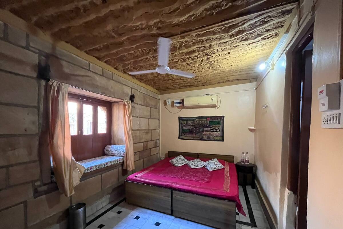 A small, rustic room with a single bed, a window with curtains, a ceiling fan, and a wall-mounted air conditioner.