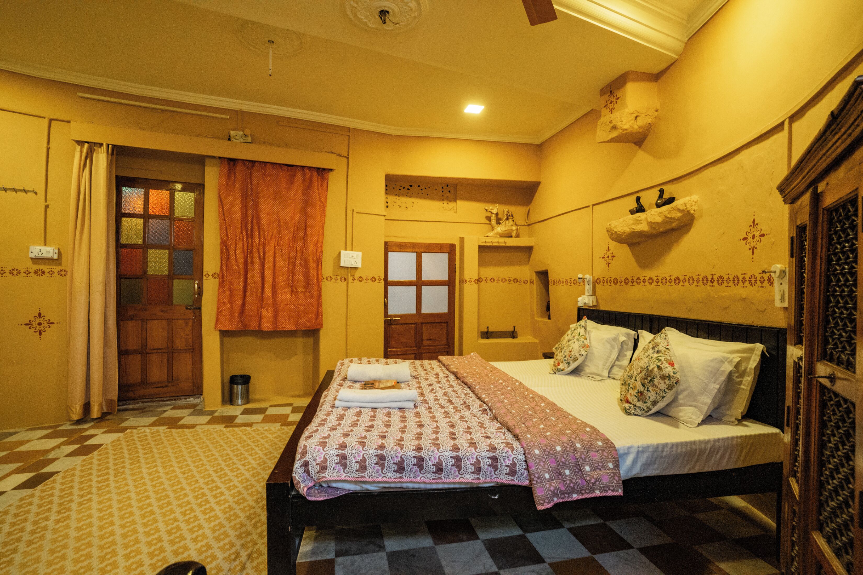 A hotel room with a checkered floor, a bed with patterned bedding, a window with curtains, and a wall-mounted shelf with decorative items.