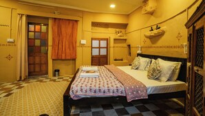 Una habitación de hotel con piso a cuadros, una cama con ropa de cama estampada, un ventanal con cortinas y un estante montado en la pared con adornos.