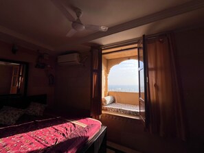 A bedroom with a bed, an open window, a ceiling fan, and an air conditioning unit. - Hotel Surja Jaisalmer (Jaisalmer)