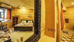 Una habitación de hotel con piso a cuadros, una cama con ropa de cama floral, una mecedora de madera, una ventana con cortinas y un espejo que refleja parte de la habitación.