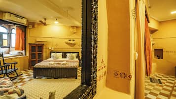 A hotel room with a checkered floor, a bed with floral bedding, a wooden rocking chair, a window with curtains, and a mirror reflecting part of the room.