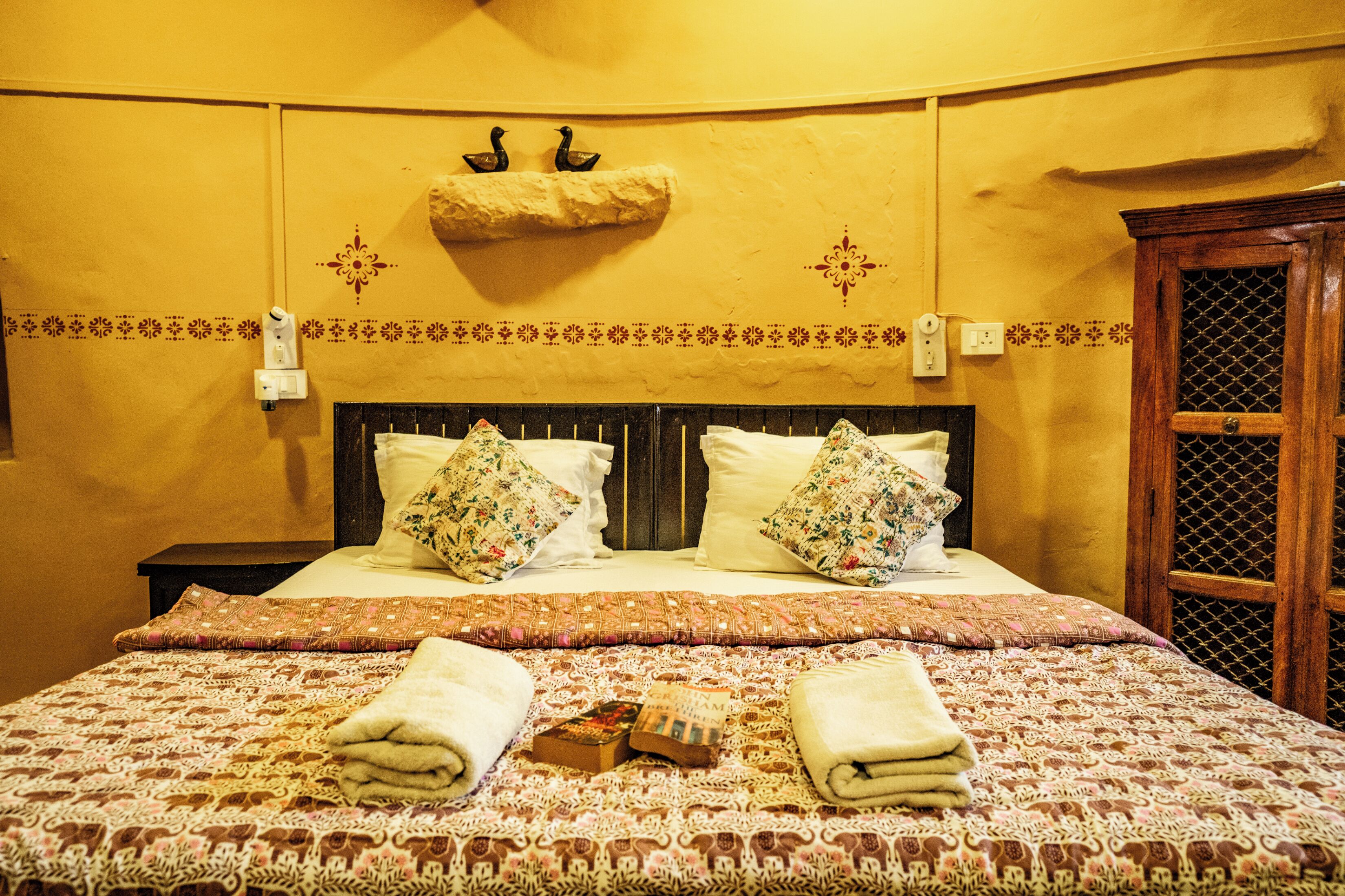 A hotel room with a bed, two pillows, a bedside table, a nightstand, a wall-mounted light switch, a decorative wall hanging, and a window with a wooden frame.