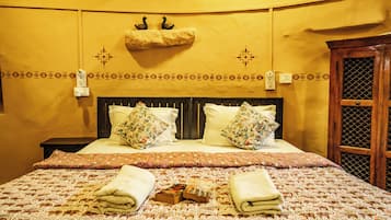 A hotel room with a bed, two pillows, a bedside table, a nightstand, a wall-mounted light switch, a decorative wall hanging, and a window with a wooden frame.