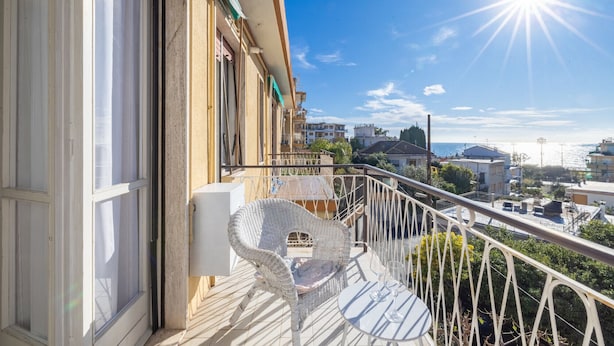Un balcón con una silla de mimbre blanca y una mesita pequeña, con vistas a un paisaje urbano de edificios y cielo despejado.