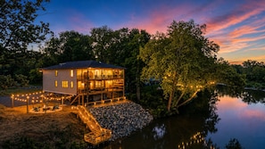 Une maison à deux étages avec une terrasse, un foyer extérieur et un mur de pierres, située au bord d’un lac.