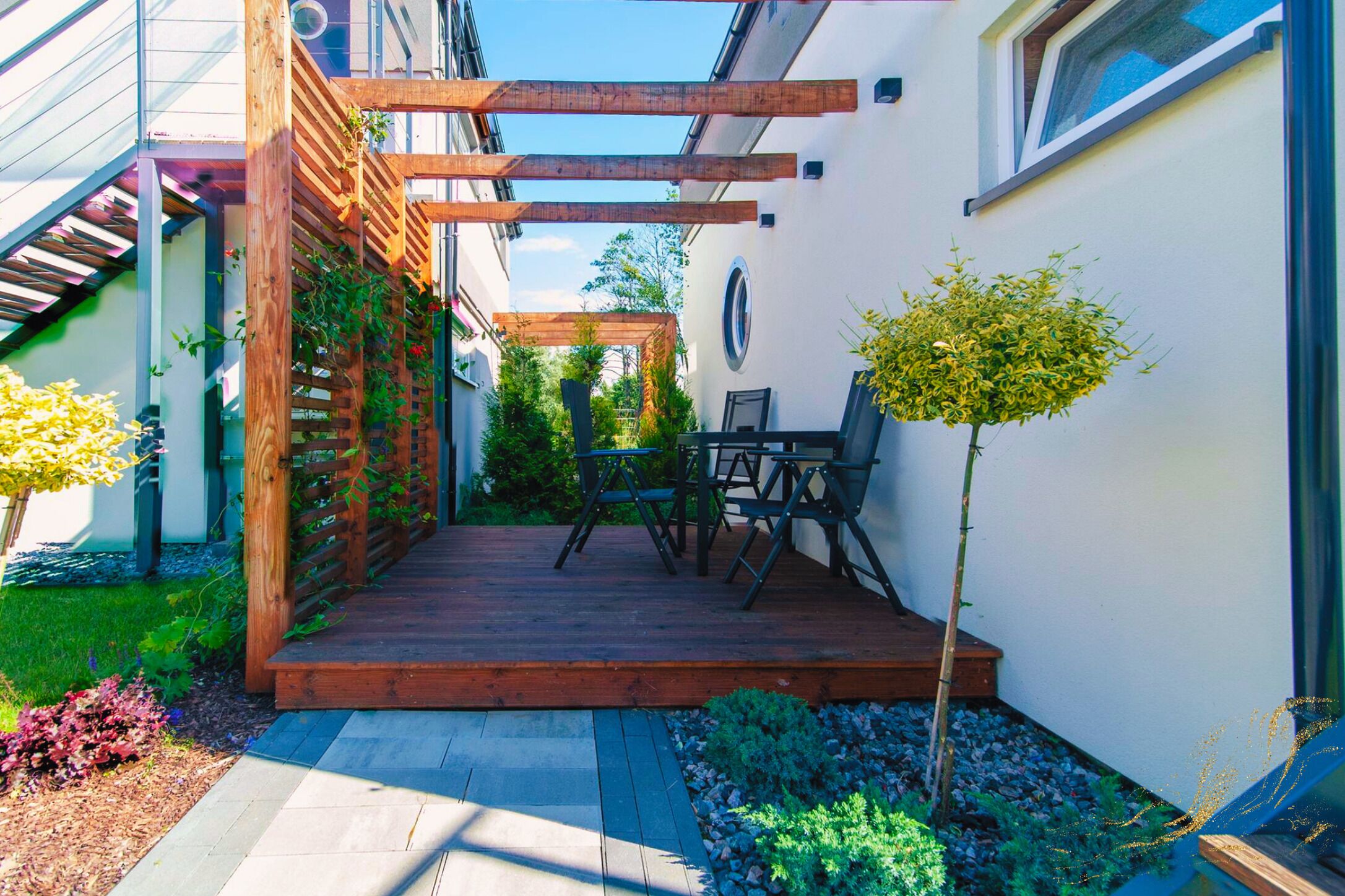 Una terraza de madera con una pérgola, una mesita y sillas, y una planta en maceta.