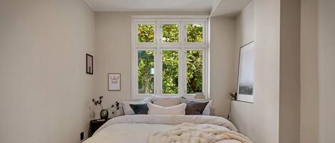 A bedroom with a large bed, a window with a view of greenery, a bedside table with a vase, and a framed picture on the wall.