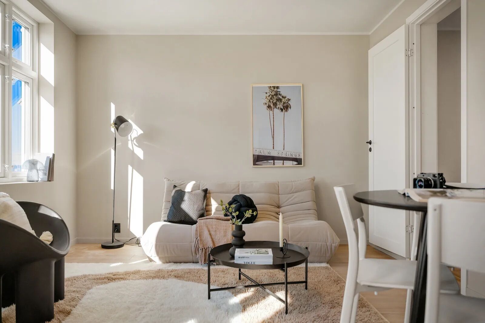 A modern living room with a sofa, a coffee table, and a lamp. There is a framed picture on the wall and a door to the right.