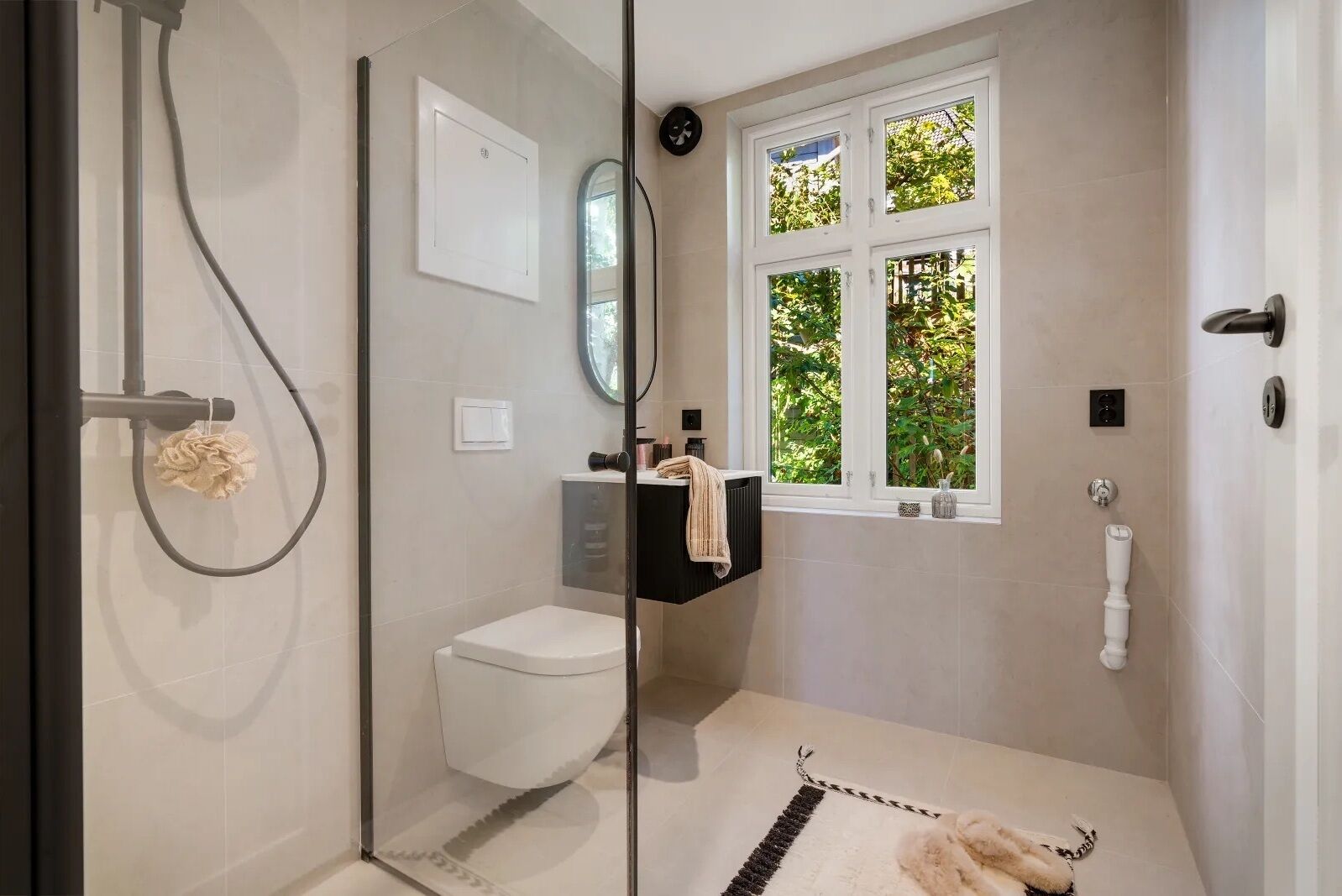 A modern bathroom with a glass shower enclosure, a toilet, a sink with a towel, and a window with a view of greenery.