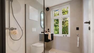 A modern bathroom with a glass shower enclosure, a toilet, a sink with a towel, and a window with a view of greenery.