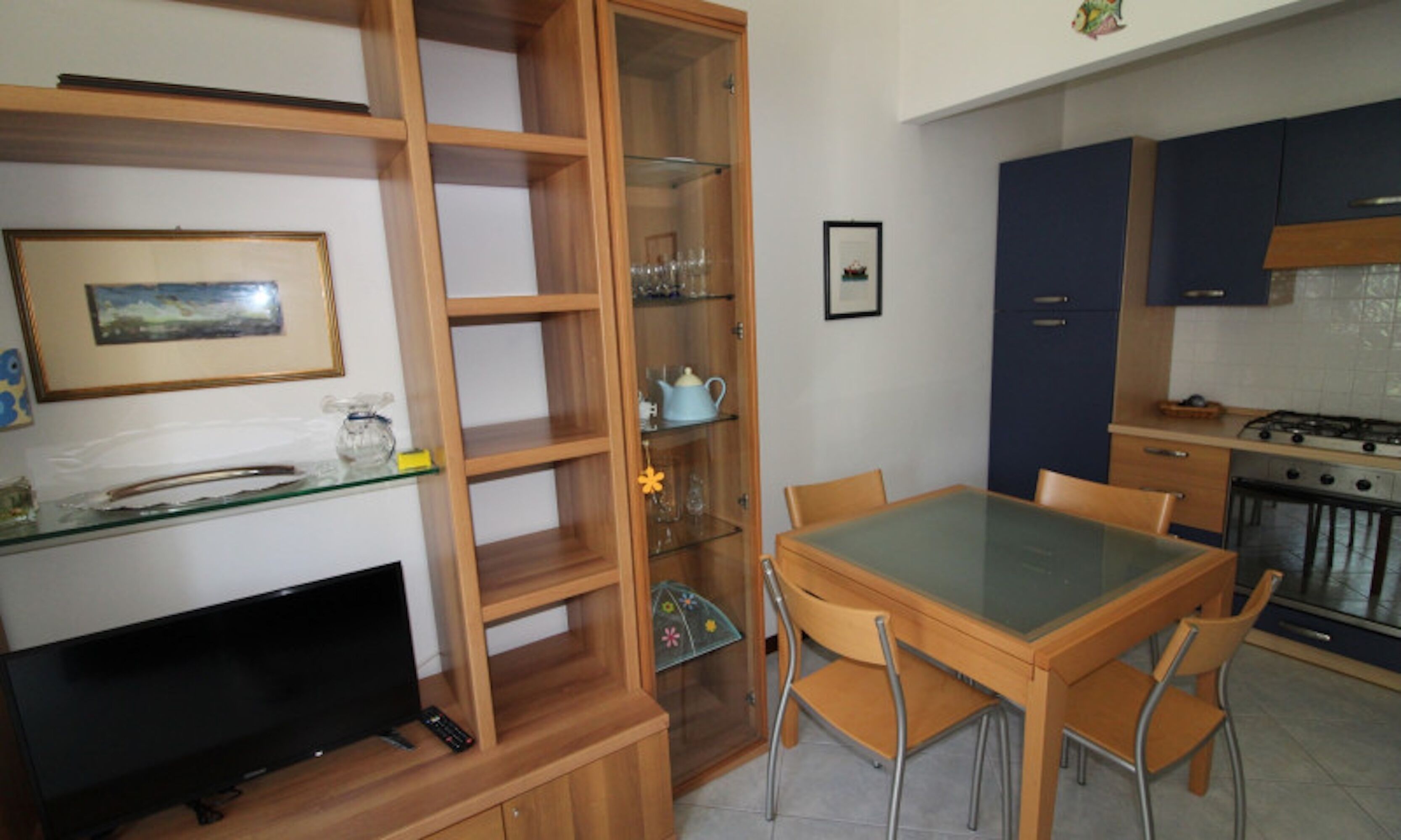 Un ambiente compatto con TV, scaffali in legno, un tavolo da pranzo con sedie e angolo cottura con pensili e fornello.