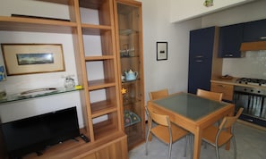 A compact living space with a TV, wooden shelving, a dining table with chairs, and a kitchen area with cabinets and a stove. - Villa 500mt From the Beach (Lignano Sabbiadoro)