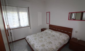 Quarto com cama, mesa de cabeceira, cômoda, janela com persianas e espelho na parede.