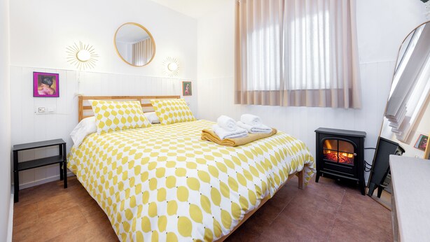 A bedroom with a bed, a small table, a mirror, a wall-mounted heater, and a window with curtains. - Tropical Garden (Lower 2) (Granada)