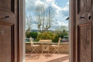 Eine offene Holztür führt auf eine Terrasse mit Tisch und Stühlen, mit Blick auf Bäume und Hügel, und einem wolkenlosen Himmel.