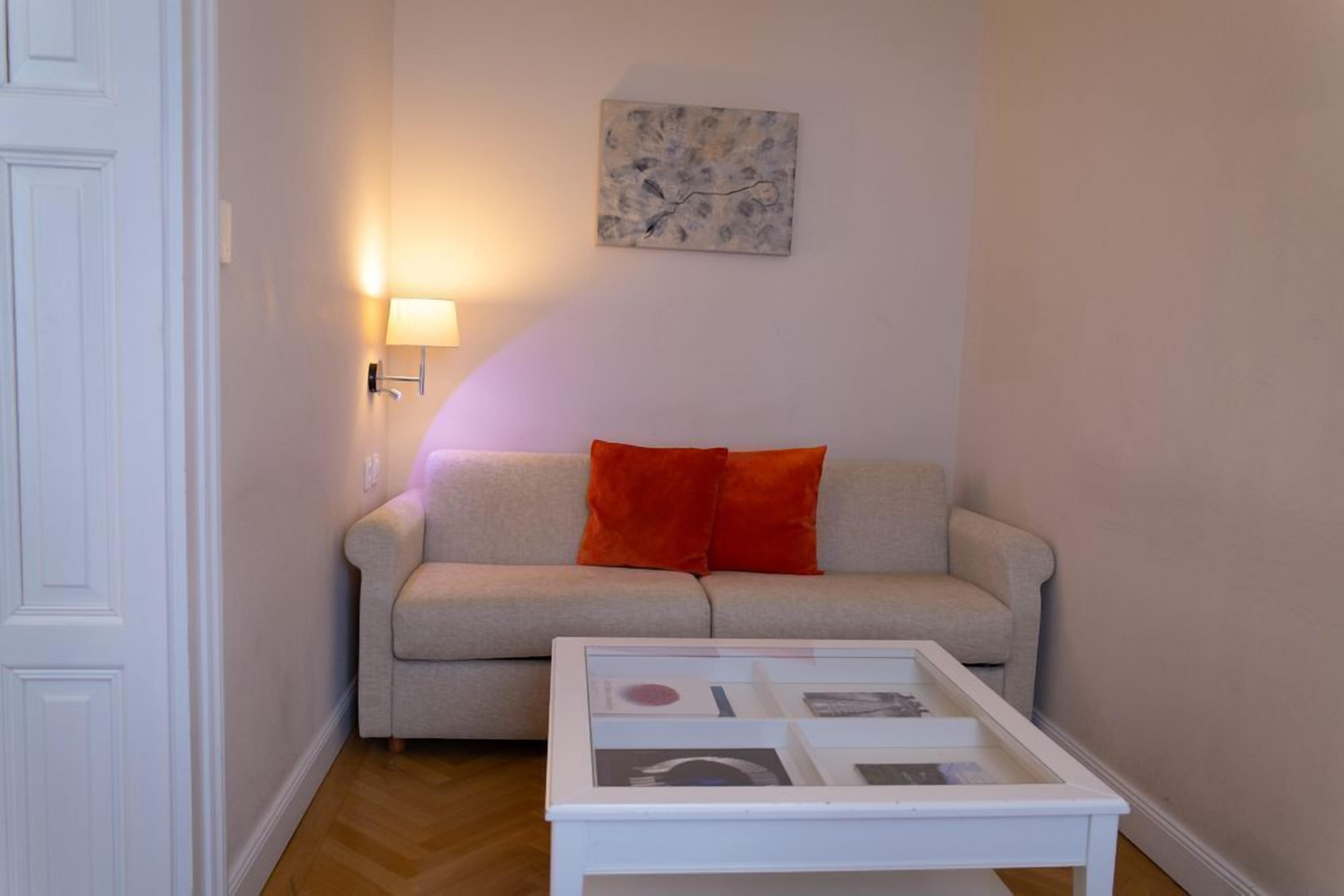 Uma sala de estar com sofá, mesa de centro de vidro, luminária na parede e uma obra de arte emoldurada.