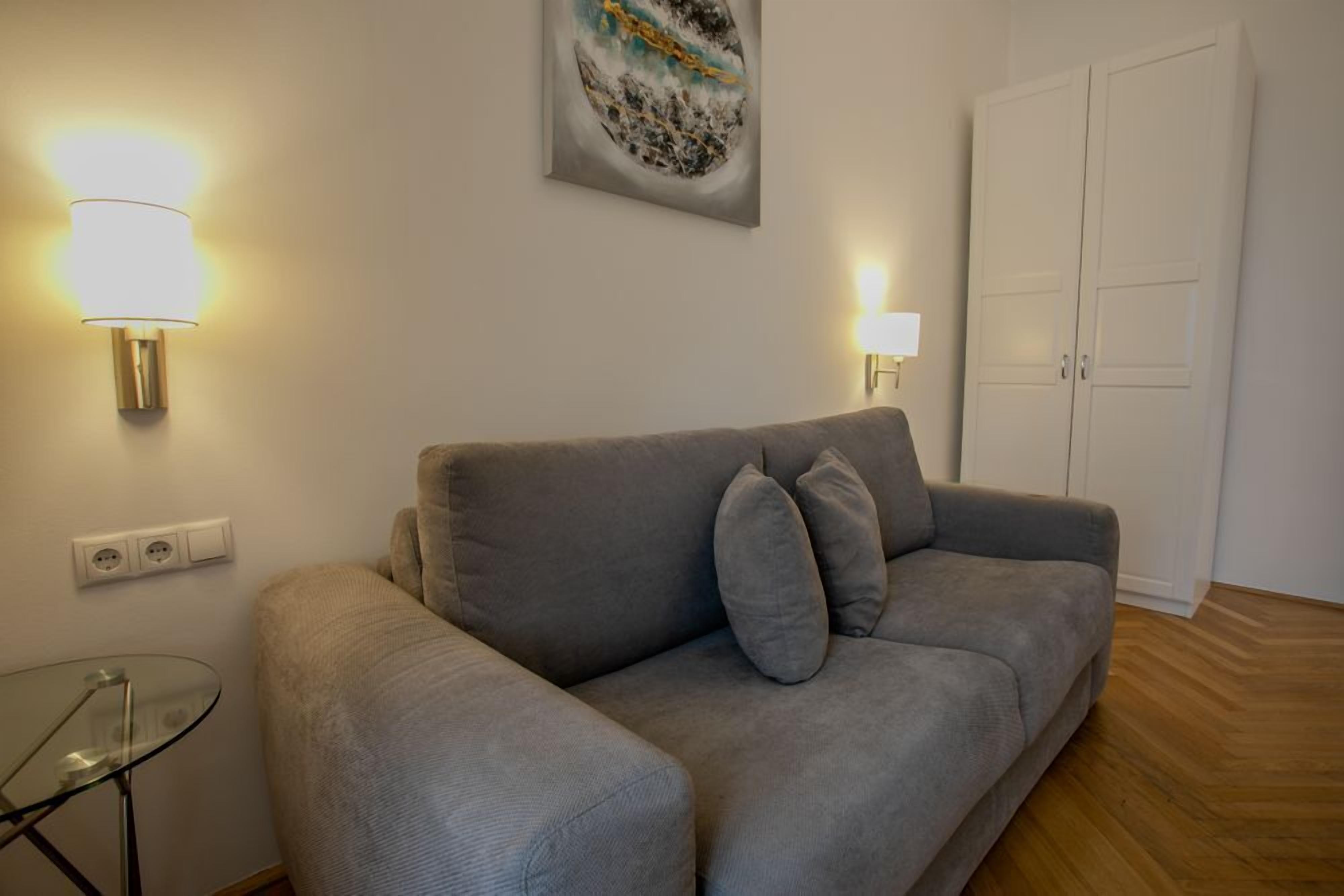 A grey sofa with two cushions, a glass table, two wall-mounted lamps, a painting, and a white wardrobe.