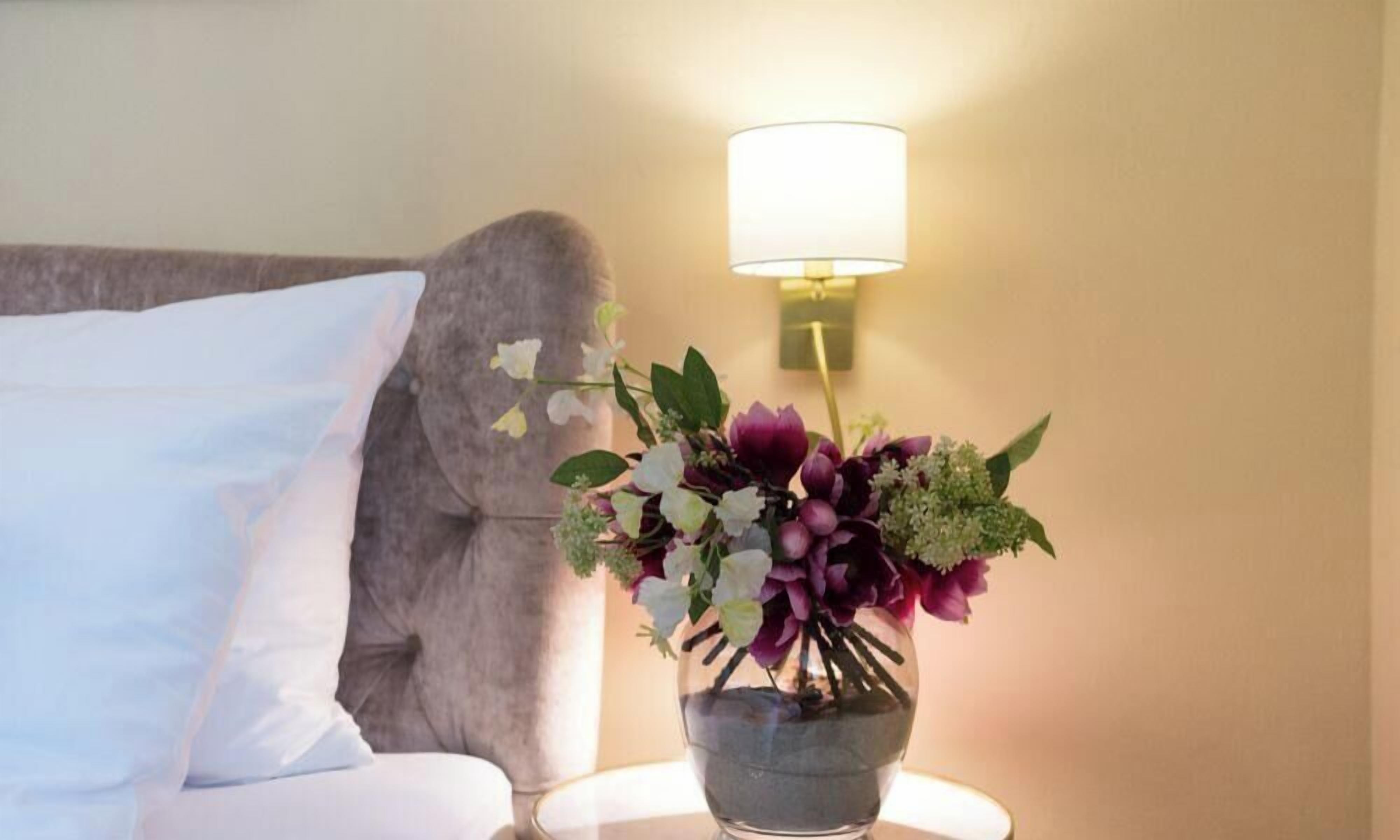 A bed with a white pillow, a grey headboard, and a bedside table with a clear vase of flowers.
