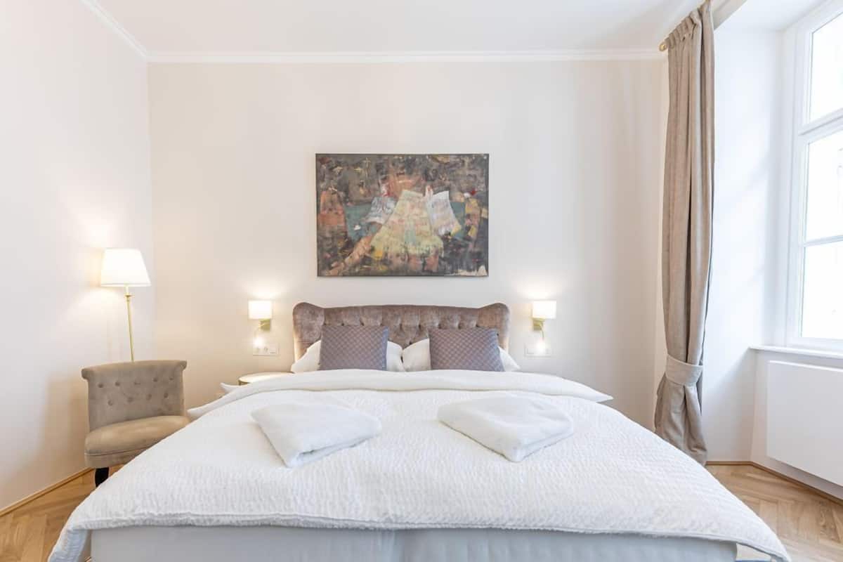 Ein Schlafzimmer mit einem großen Bett, zwei Nachttischlampen, einem Sessel und einem Bild an der Wand.