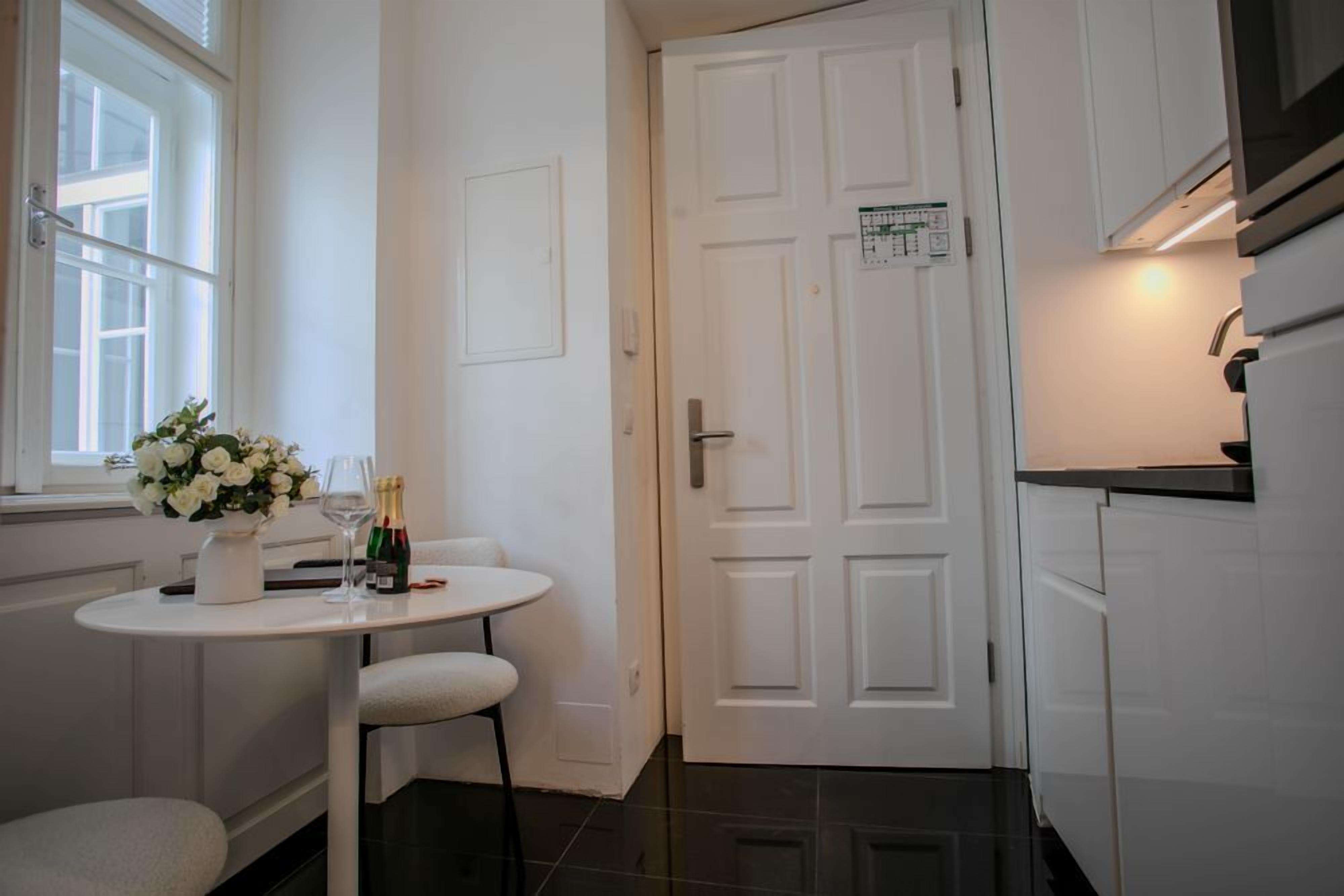 A kitchen with white cabinets, a white door with a calendar, a small round table with a vase of flowers, and a bottle of champagne.