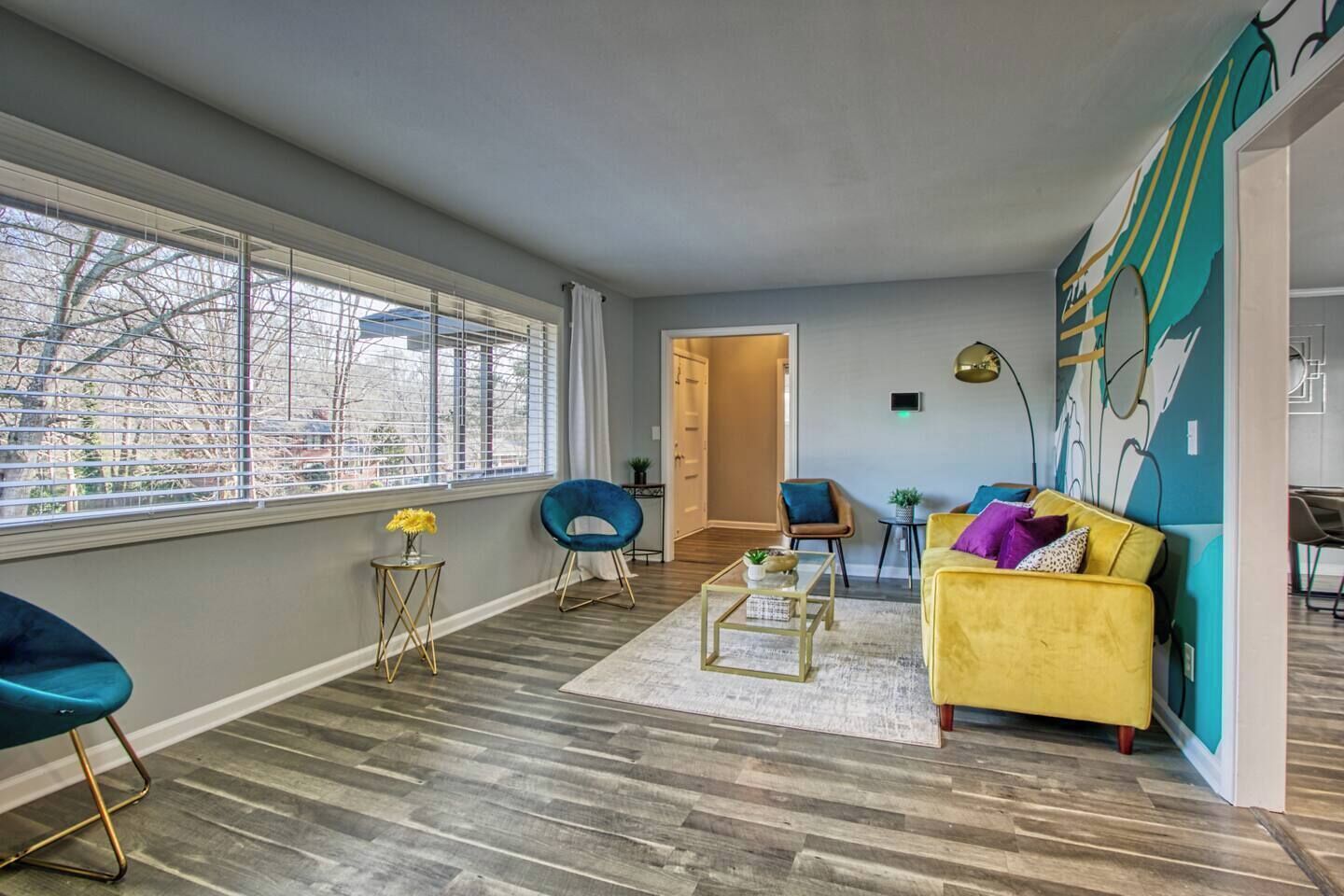 A modern living room with a yellow sofa, blue chairs, and a large window with blinds.