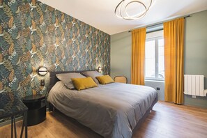 Un dormitorio moderno con papel tapiz estampado, una cama con ropa de cama gris, una mesa redonda y una ventana con cortinas.