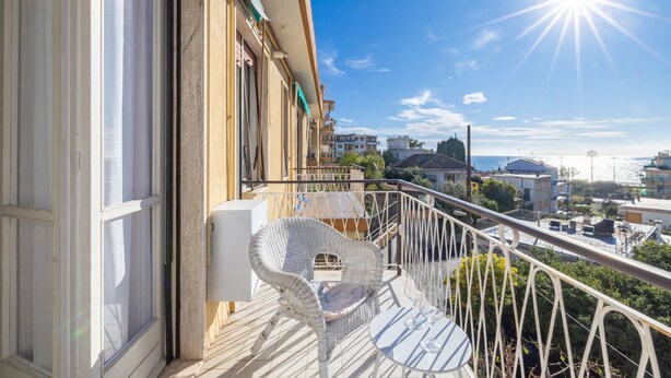 Un balcón con una silla de mimbre blanca y una mesita pequeña, con vistas a un paisaje urbano de edificios y cielo despejado.