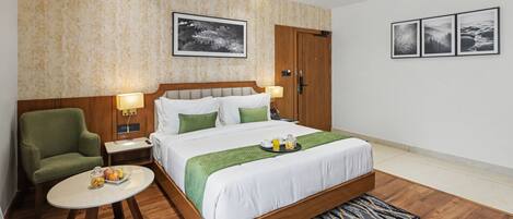 A hotel room with a bed, a chair, a small table with fruit, a nightstand, a lamp, and a wall with three framed pictures.